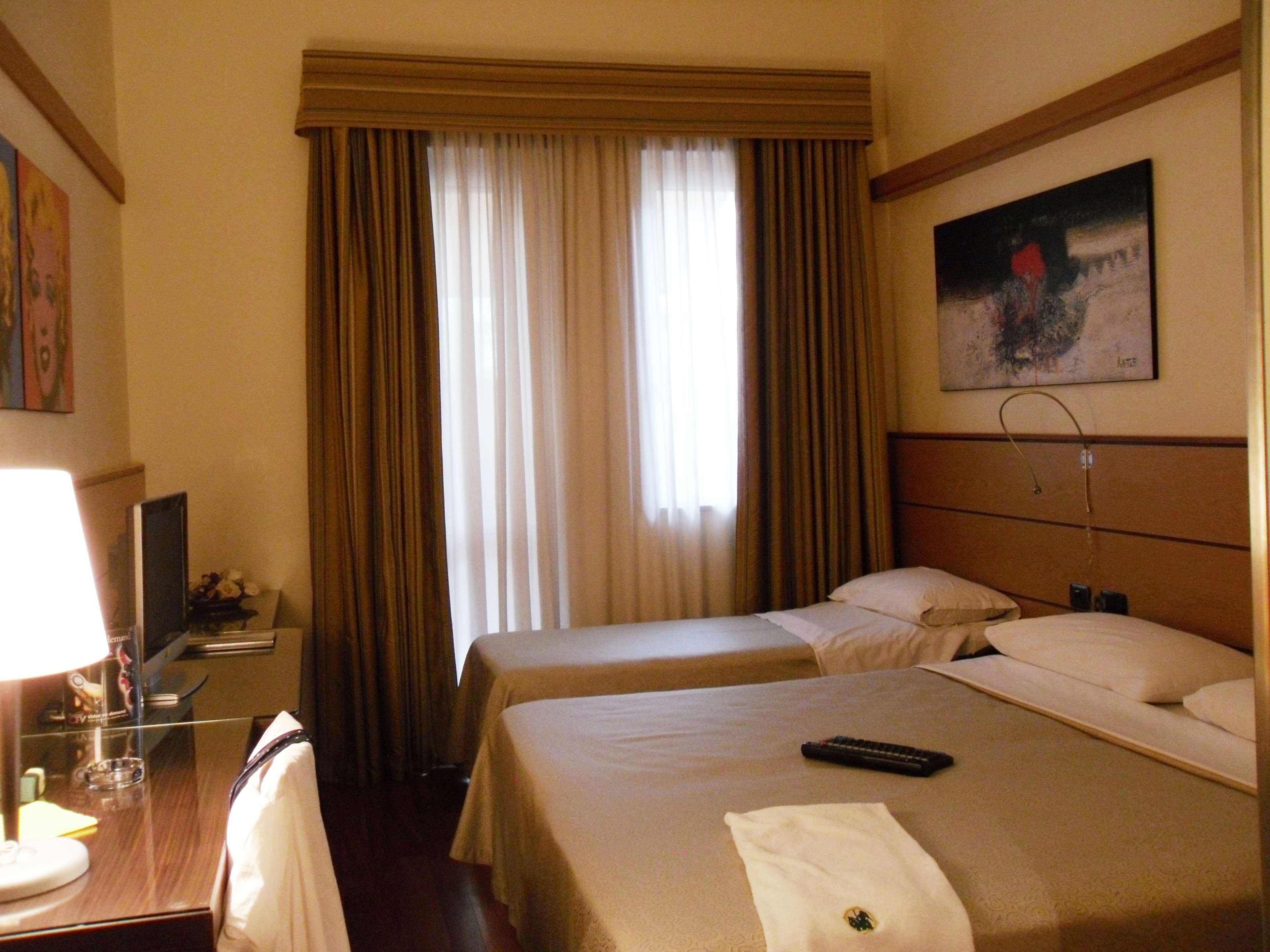 A hotel room with two beds, a desk with a lamp, a painting on the wall, and a window with curtains.