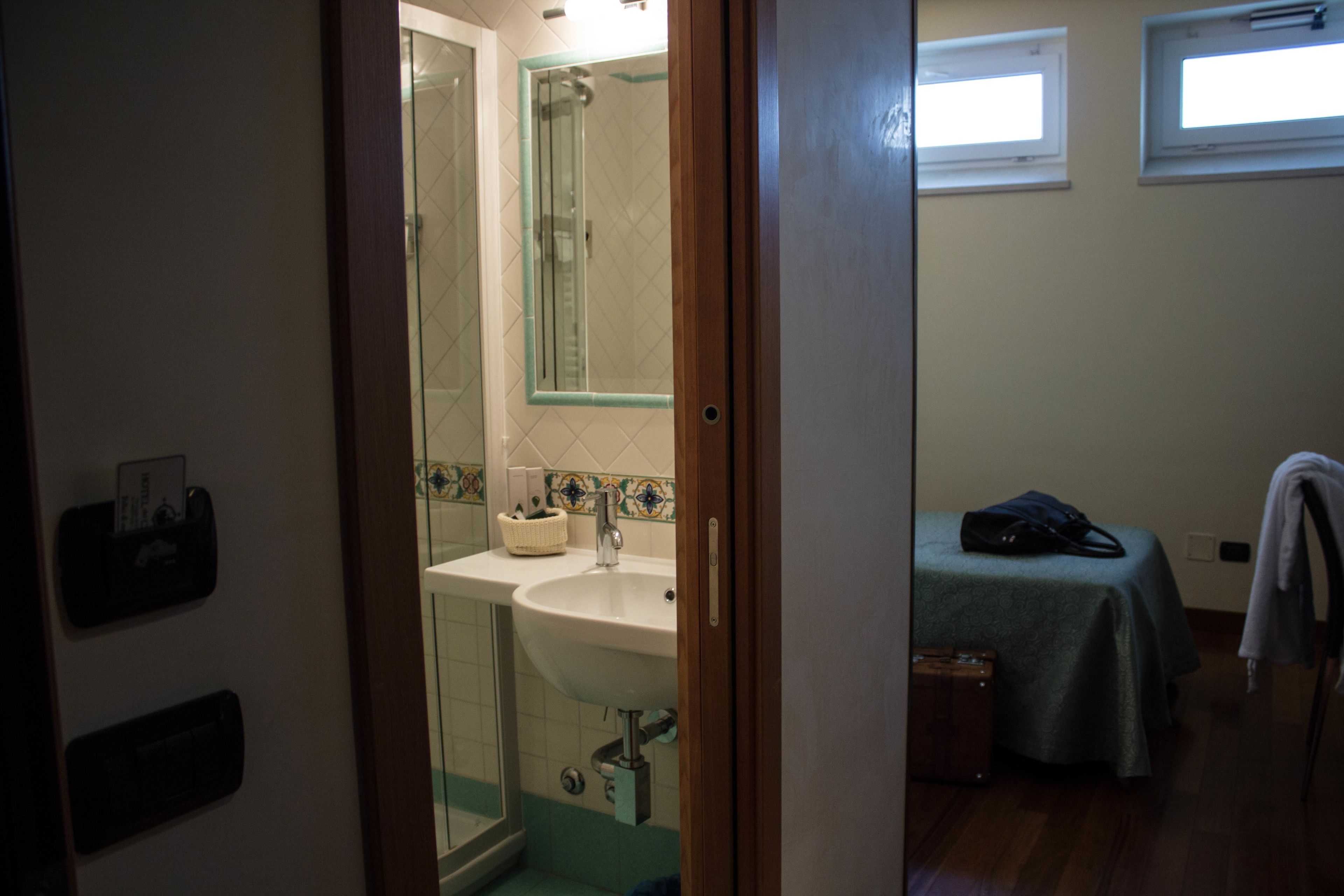 A bathroom with a sink, mirror, and shower. There is a bed with a blanket and a suitcase in the room.