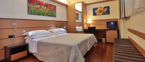 A hotel room with a bed, bedside tables, a desk with a chair, a television, and artwork on the walls.