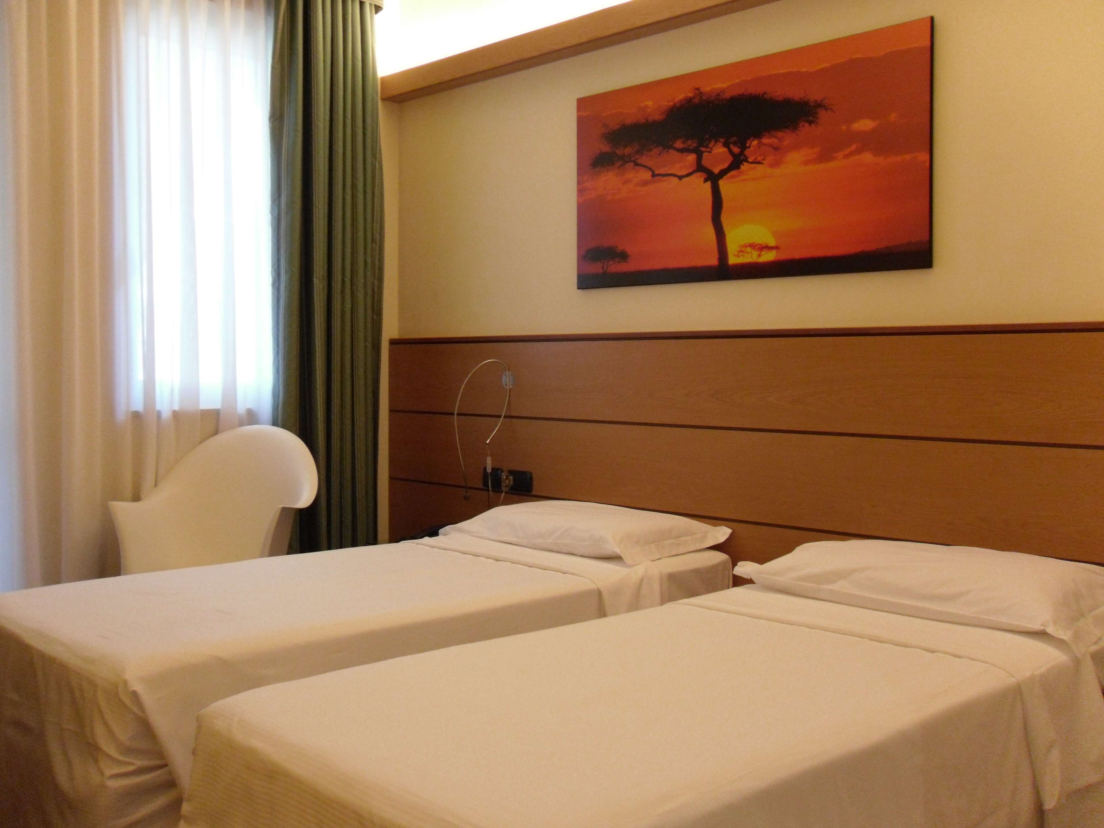 Habitación de hotel con dos camas individuales, un cabecero de madera, un cuadro de un árbol al atardecer, una silla y una ventana con cortinas.
