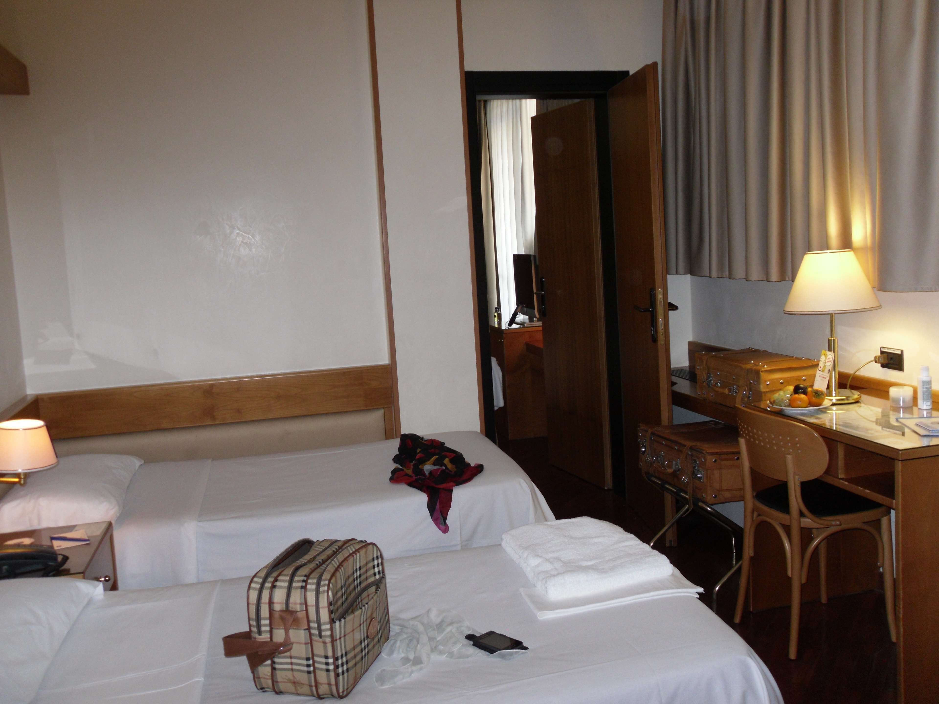 A hotel room with two beds, a desk with a lamp, a chair, and a small table with a fruit bowl.
