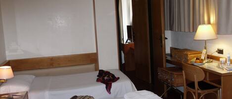 Ein Hotelzimmer mit zwei Betten, einem Schreibtisch mit Lampe, einem Sessel und einem kleinen Tisch mit Obstschale.