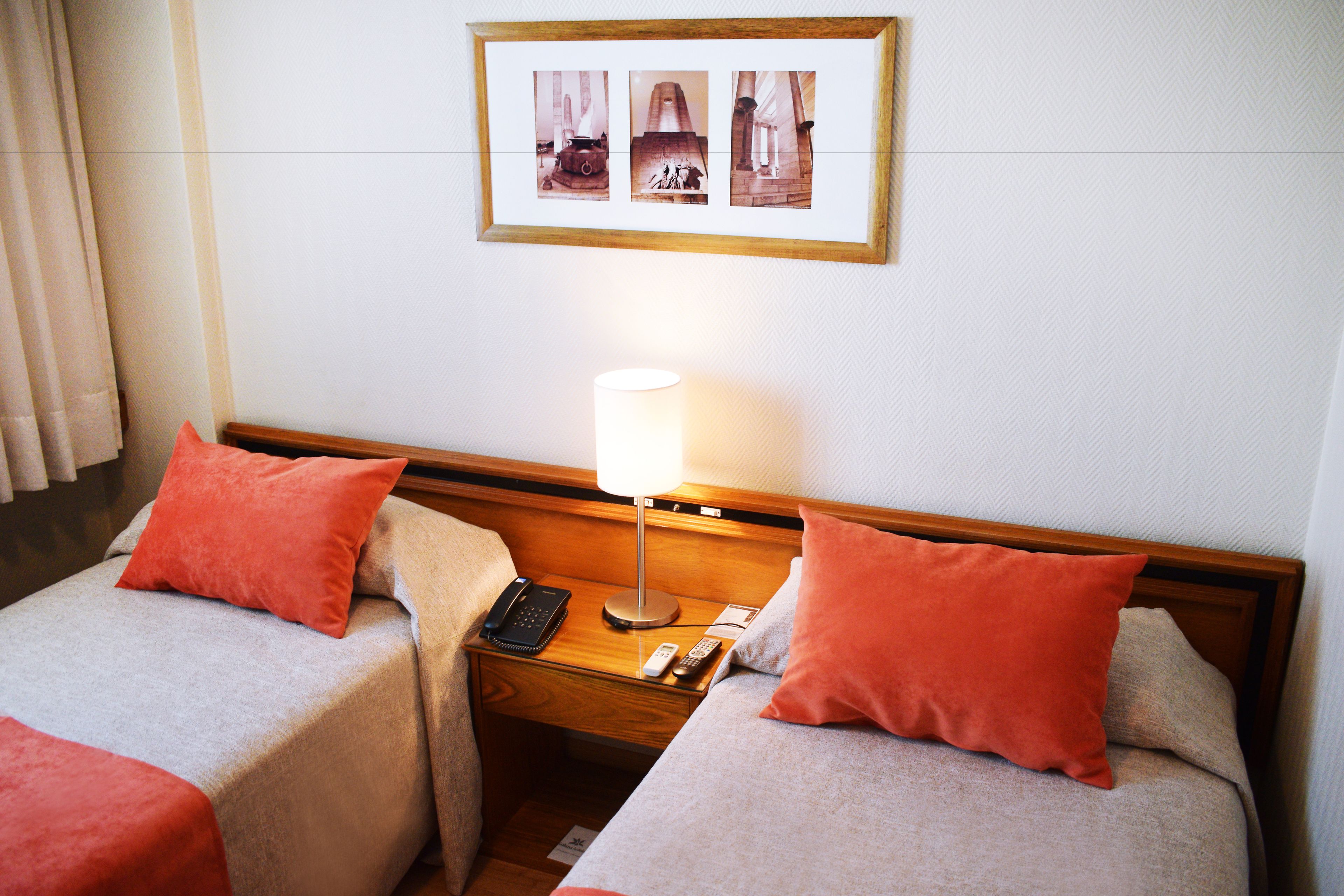 A hotel room with two beds, each with a red pillow, a bedside table with a lamp, a phone, and a remote control.