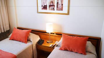 A hotel room with two beds, each with a red pillow, a bedside table with a lamp, a phone, and a remote control.