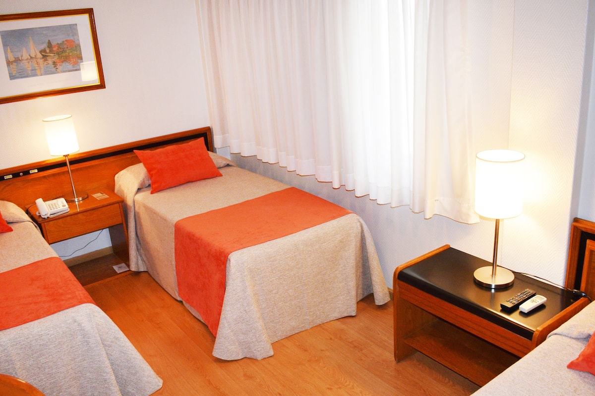A hotel room with two single beds, a wooden nightstand with a lamp, a telephone, and a remote control.