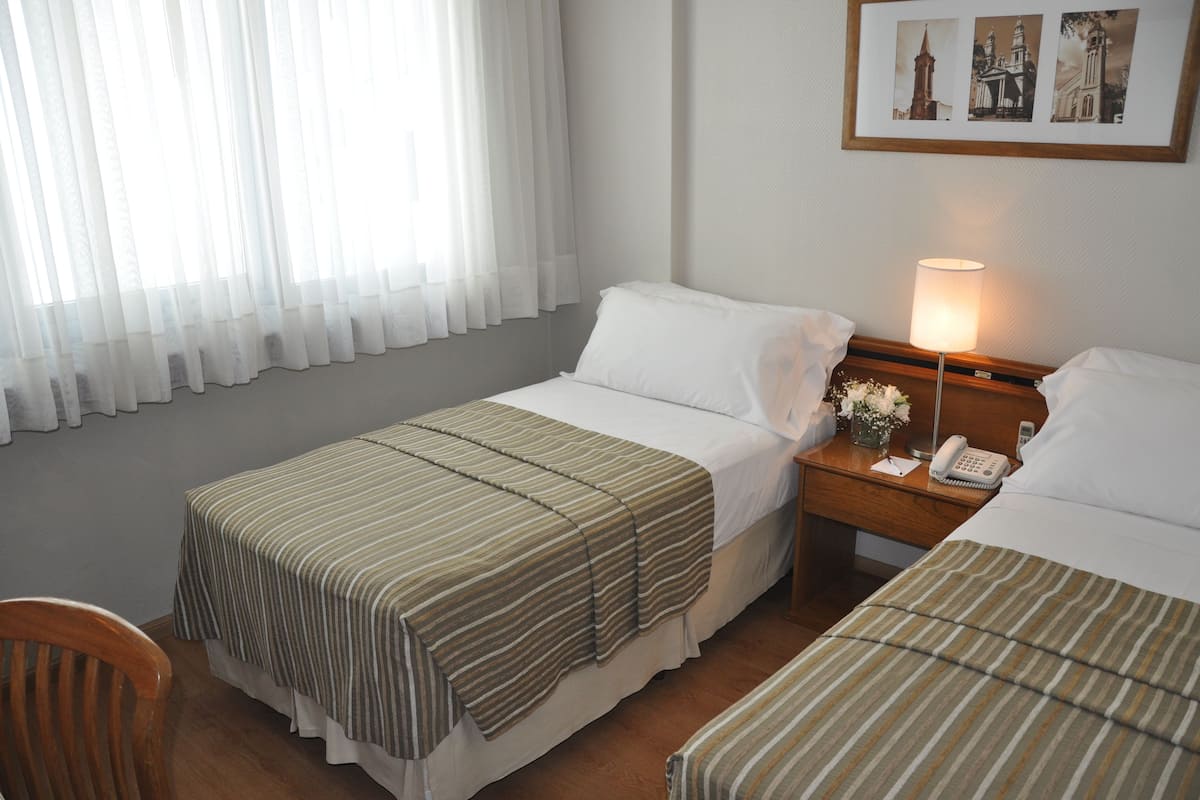 A hotel room with two beds, a wooden nightstand with a lamp, a phone, and a framed picture on the wall.