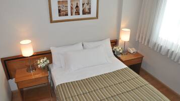 A neatly made bed with white linens, two bedside tables with lamps, a framed picture above the bed, and a window with sheer curtains.