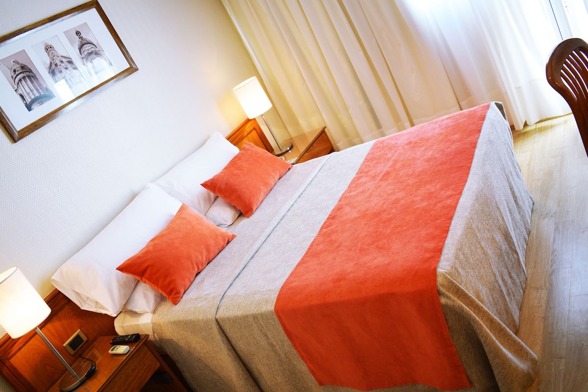 A hotel room with a bed, pillows, a blanket, a nightstand with a lamp, a remote control, and a framed picture on the wall.