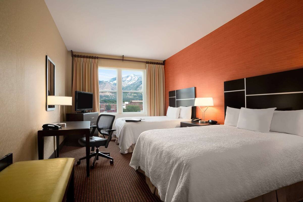 A hotel room with two beds, a desk with a chair, a TV, and a view of mountains through the window.
