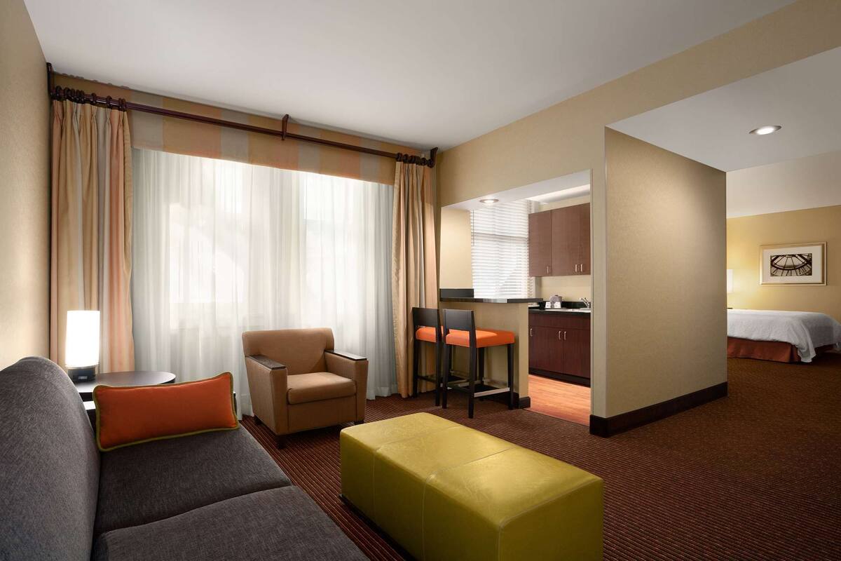 A hotel room with a sofa, ottoman, dining table, chairs, kitchenette, and a bed.