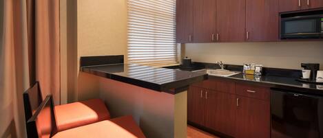 Una cocina compacta con armarios de madera oscura, encimera negra y microondas integrado. Hay dos taburetes naranja y una ventana con persianas.