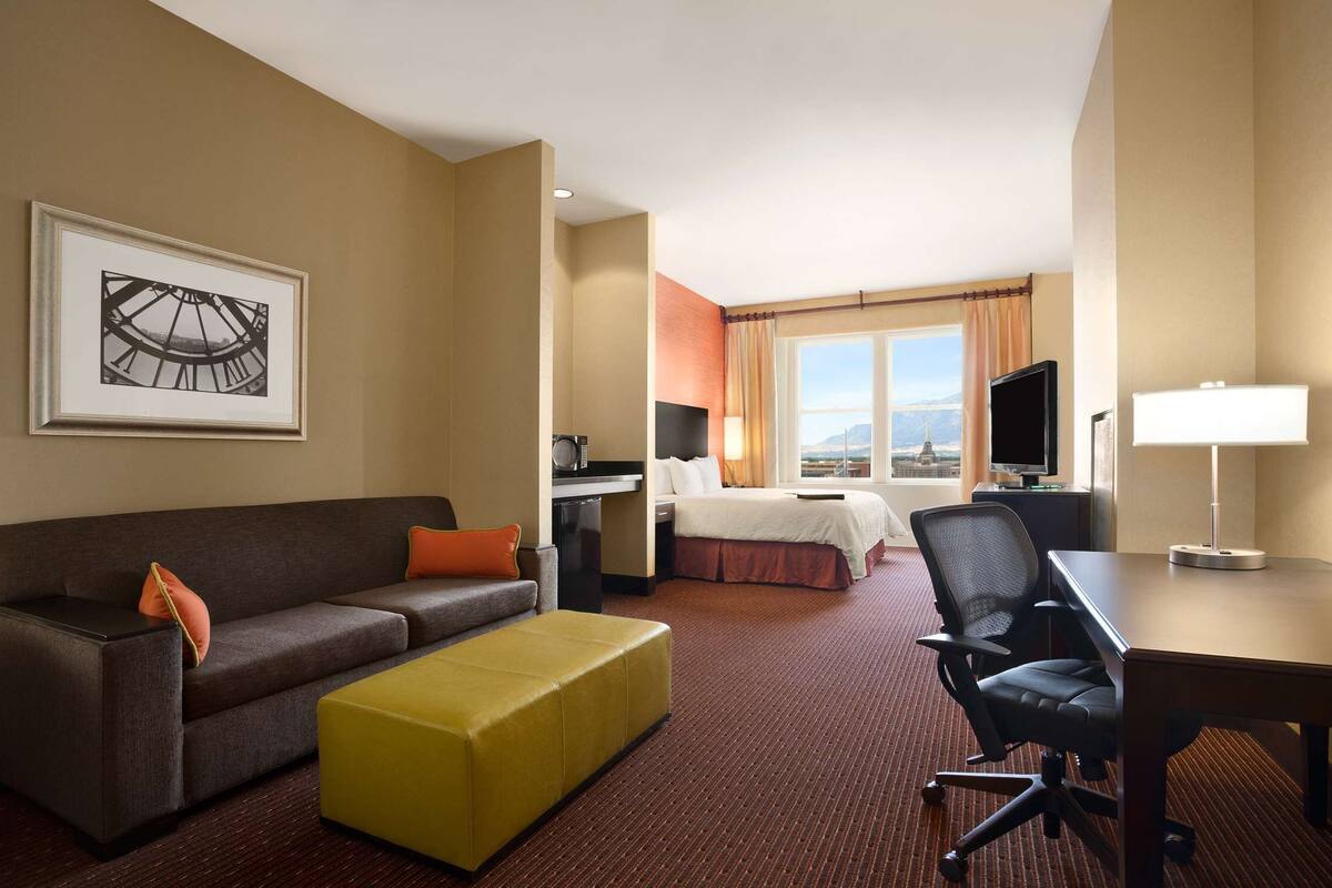 A hotel room with a sofa, ottoman, desk, chair, bed, television, and a framed picture on the wall.