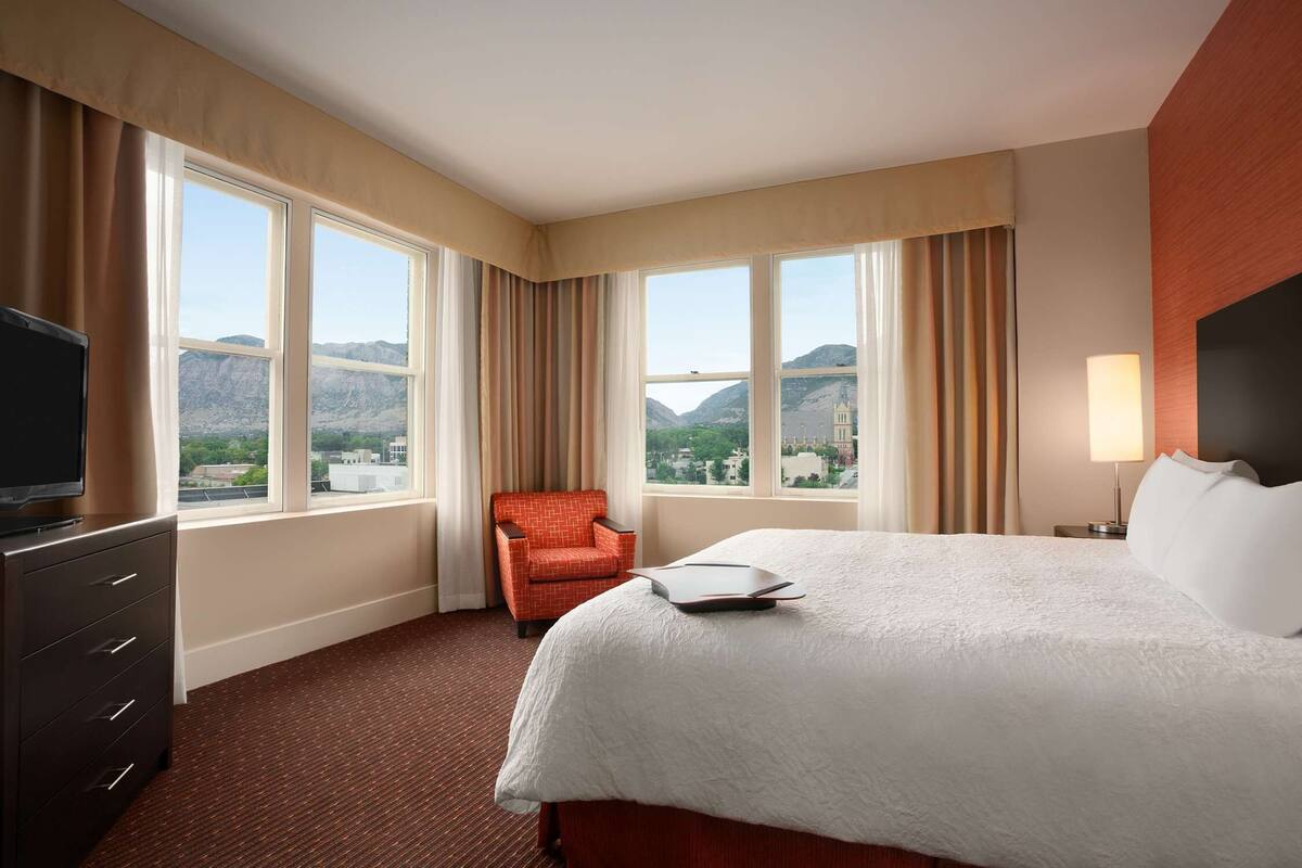 A hotel room with a large bed, a TV, a chair, and a view of mountains through the windows.