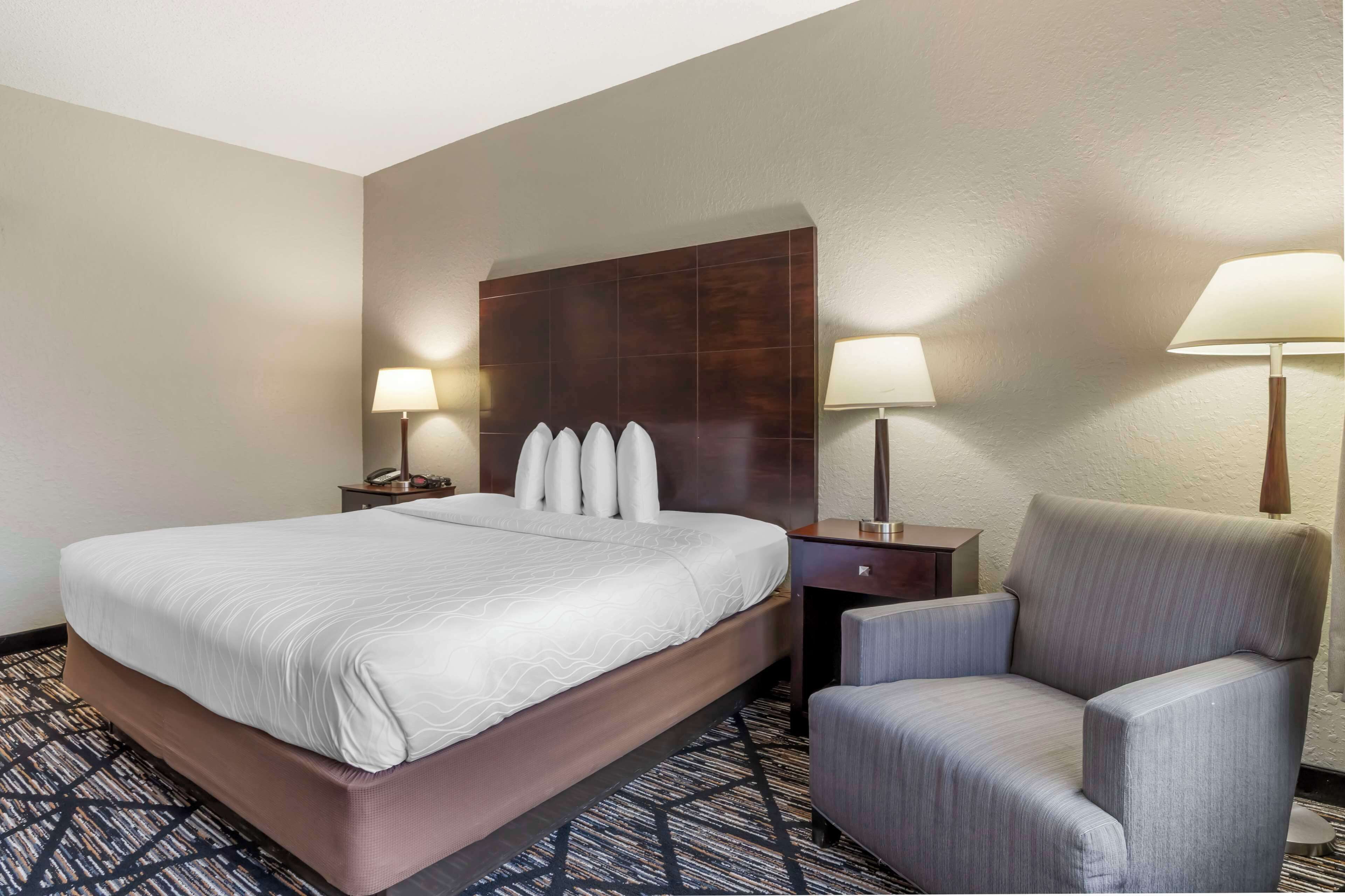 A hotel room with a large bed, two bedside tables with lamps, a chair, and a dark headboard.