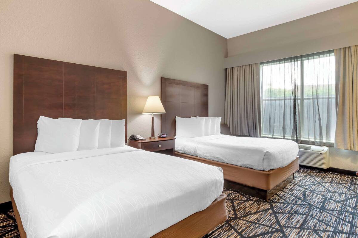 A hotel room with two beds, a nightstand, a lamp, and a large window with curtains.