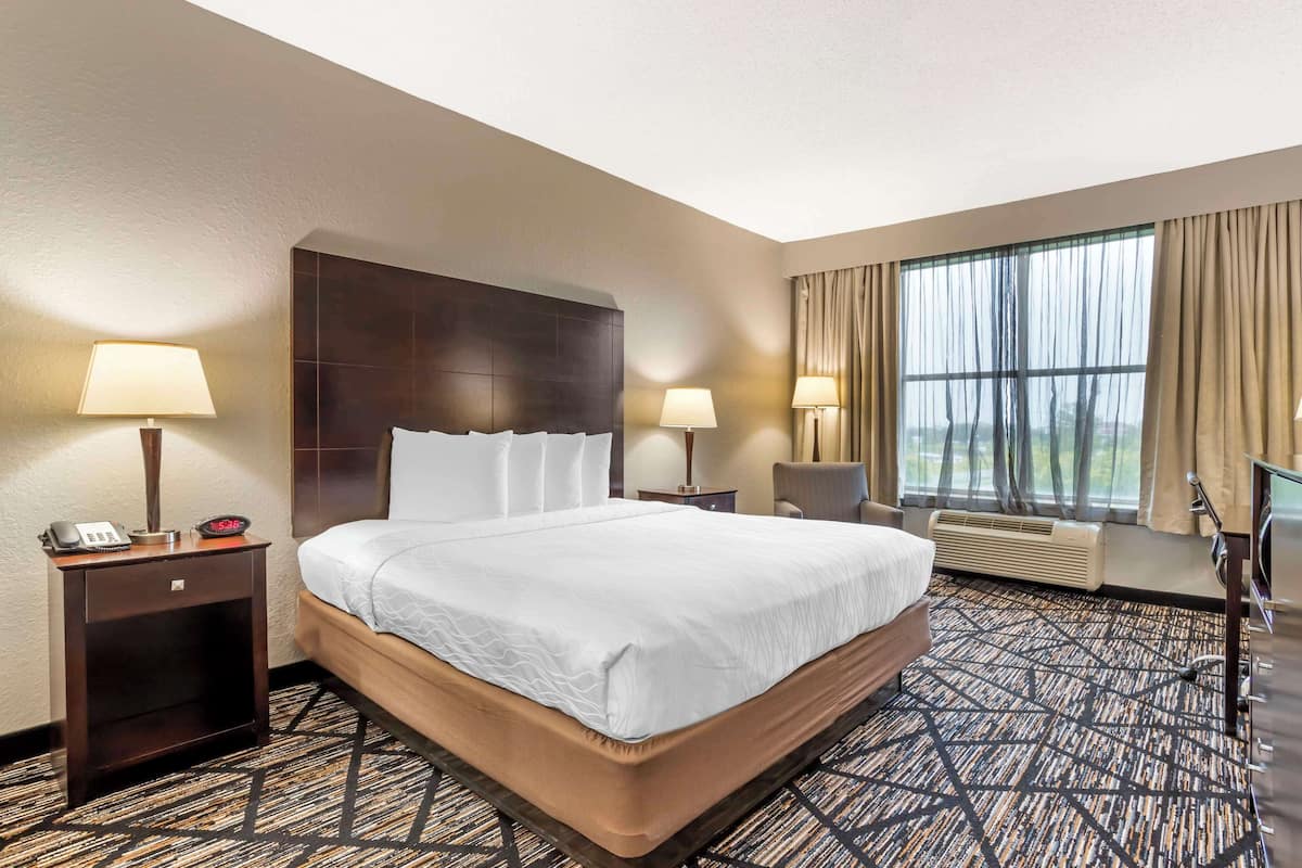 A hotel room with a large bed, two bedside tables with lamps, a chair, a desk, and a large window with curtains.