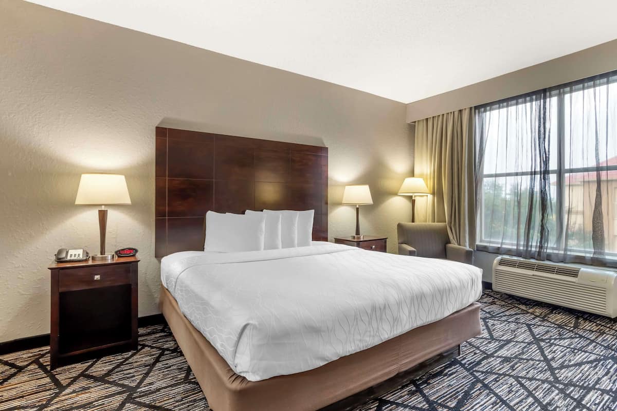 A hotel room with a large bed, two bedside tables with lamps, a chair, a window with curtains, and a wall-mounted air conditioner.