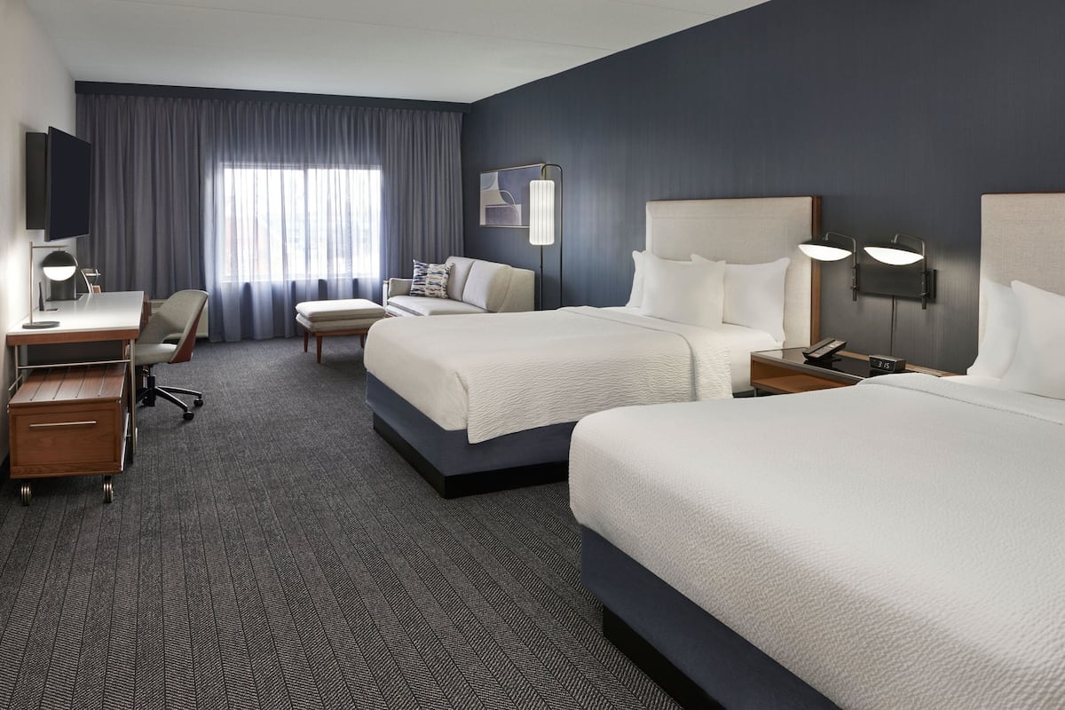 A hotel room with two beds, a desk, a chair, a TV, and a large window with curtains.