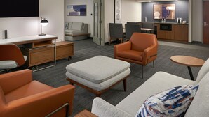 Une chambre d’hôtel moderne comprenant un canapé, un fauteuil, un pouf, un téléviseur à écran plat, un bureau avec une lampe, une kitchenette et un coin repas avec des chaises et une table.