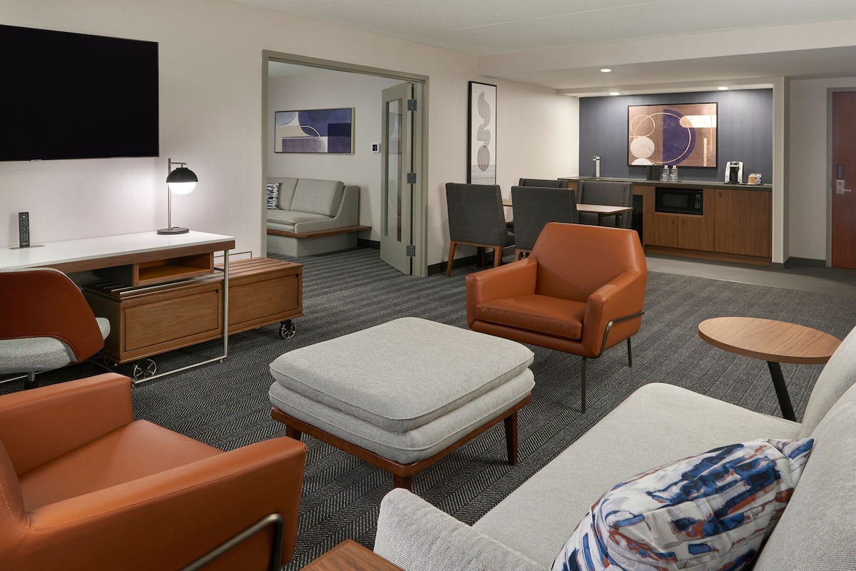 A modern hotel room with a sofa, armchair, ottoman, a flat-screen TV, a desk with a lamp, a kitchenette, and a dining area with chairs and a table.