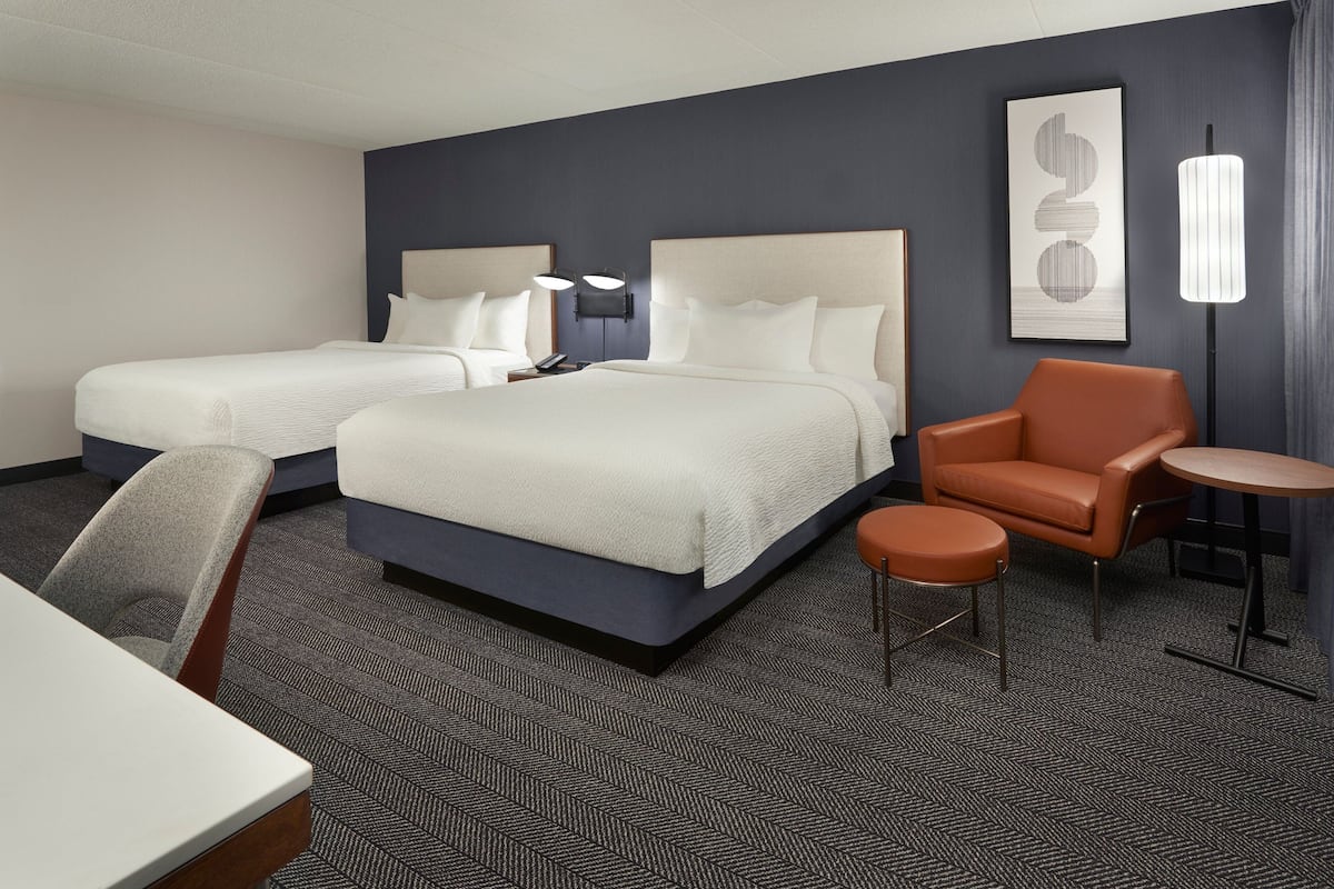 A hotel room with two beds, a desk, a chair, a round table, and a lamp.