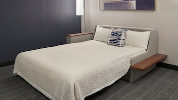 Una cama bien tendida con cubrecama blanco, una mesita de noche con lámpara y un cuadro abstracto colgado en la pared.