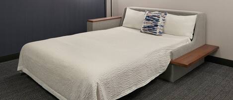 A neatly made bed with a white cover, a bedside table with a lamp, and a wall-mounted abstract painting.