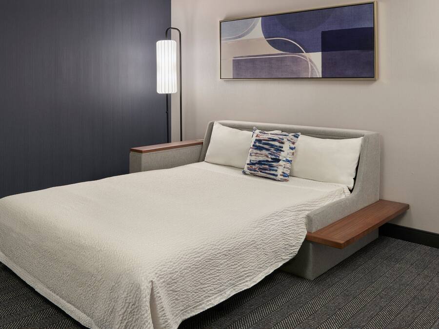 Una cama bien tendida con cubrecama blanco, una mesita de noche con lámpara y un cuadro abstracto colgado en la pared.