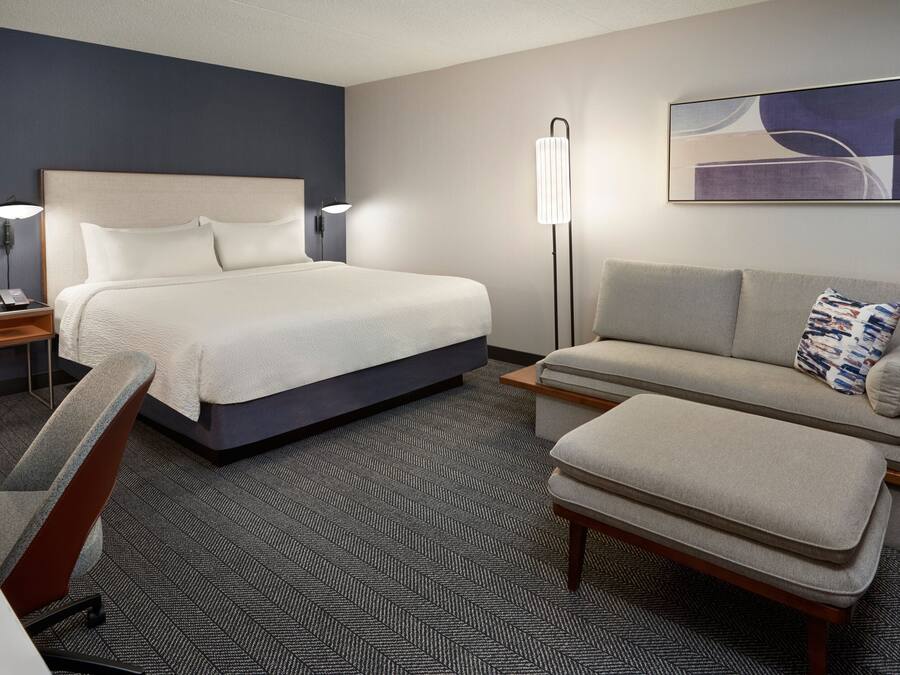 A hotel room with a bed, a sofa, a chair, a standing lamp, and a painting on the wall.