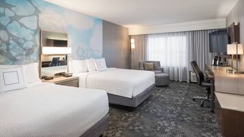 Quarto de hotel com duas camas, uma escrivaninha, uma cadeira, uma televisão e uma janela grande com cortinas.