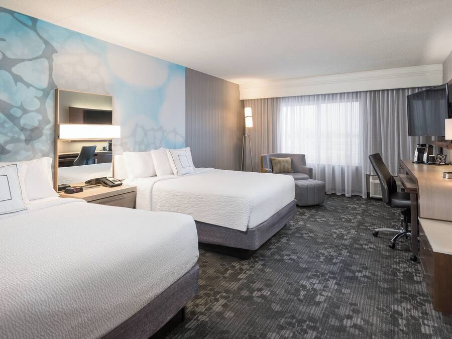 Quarto de hotel com duas camas, uma escrivaninha, uma cadeira, uma televisão e uma janela grande com cortinas.