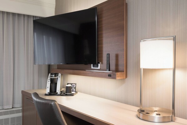 A hotel room with a flat-screen TV mounted above a wooden desk, a coffee maker, a lamp, and a chair.