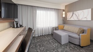 Habitación de hotel moderna con televisor de pantalla plana, sofá, escritorio con cafetera y una ventana grande con cortinas translúcidas.