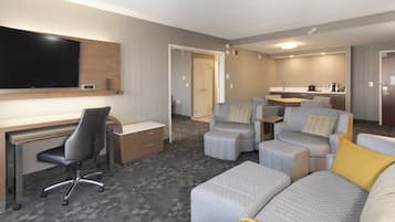 A hotel room with a flat-screen TV, a desk with a chair, a sofa, and a small kitchenette.