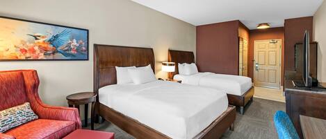 A hotel room with a large bed, two bedside tables, a desk with a chair, a red armchair, a small round table, and a painting on the wall.