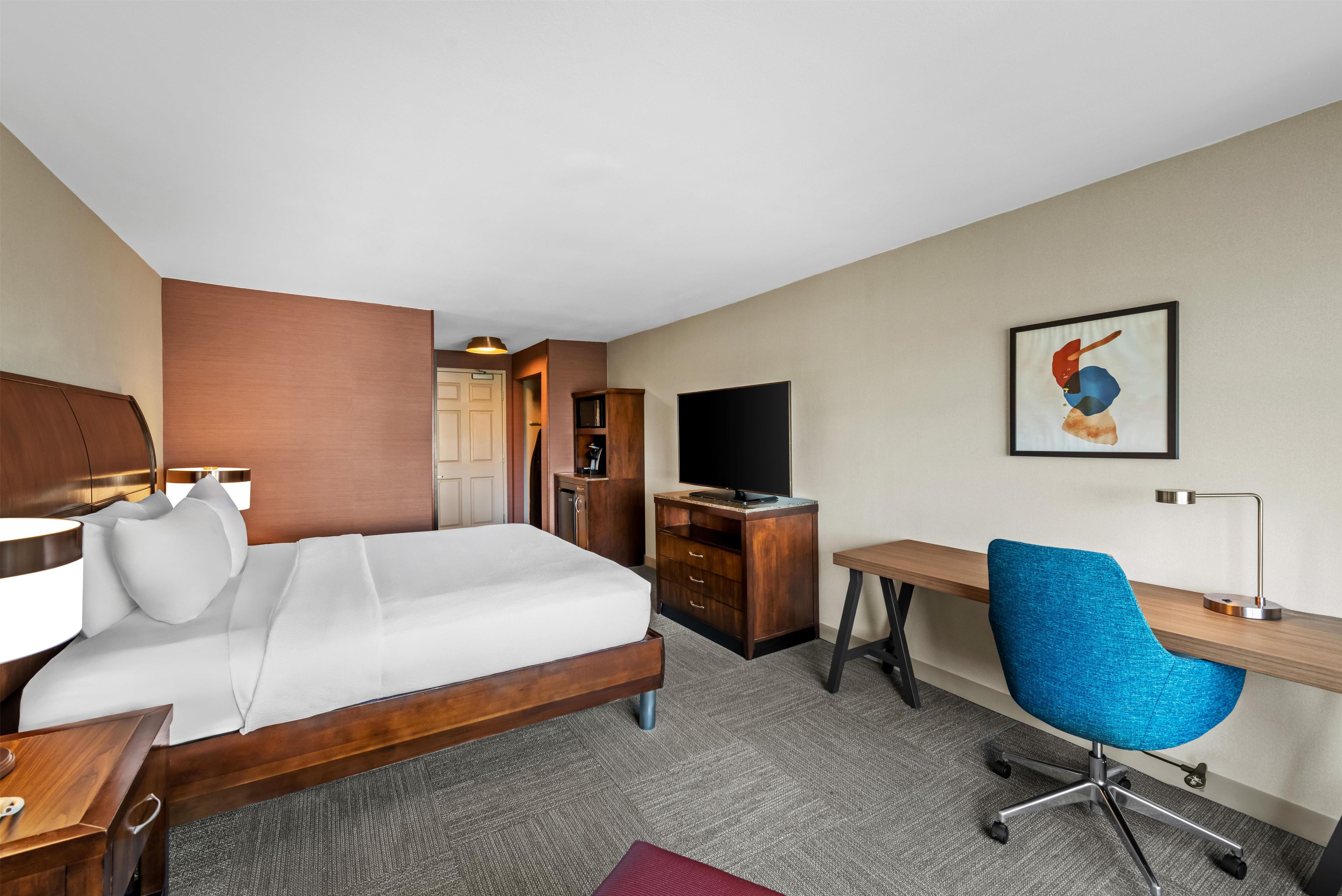 A hotel room with a bed, desk, chair, television, and a painting on the wall.
