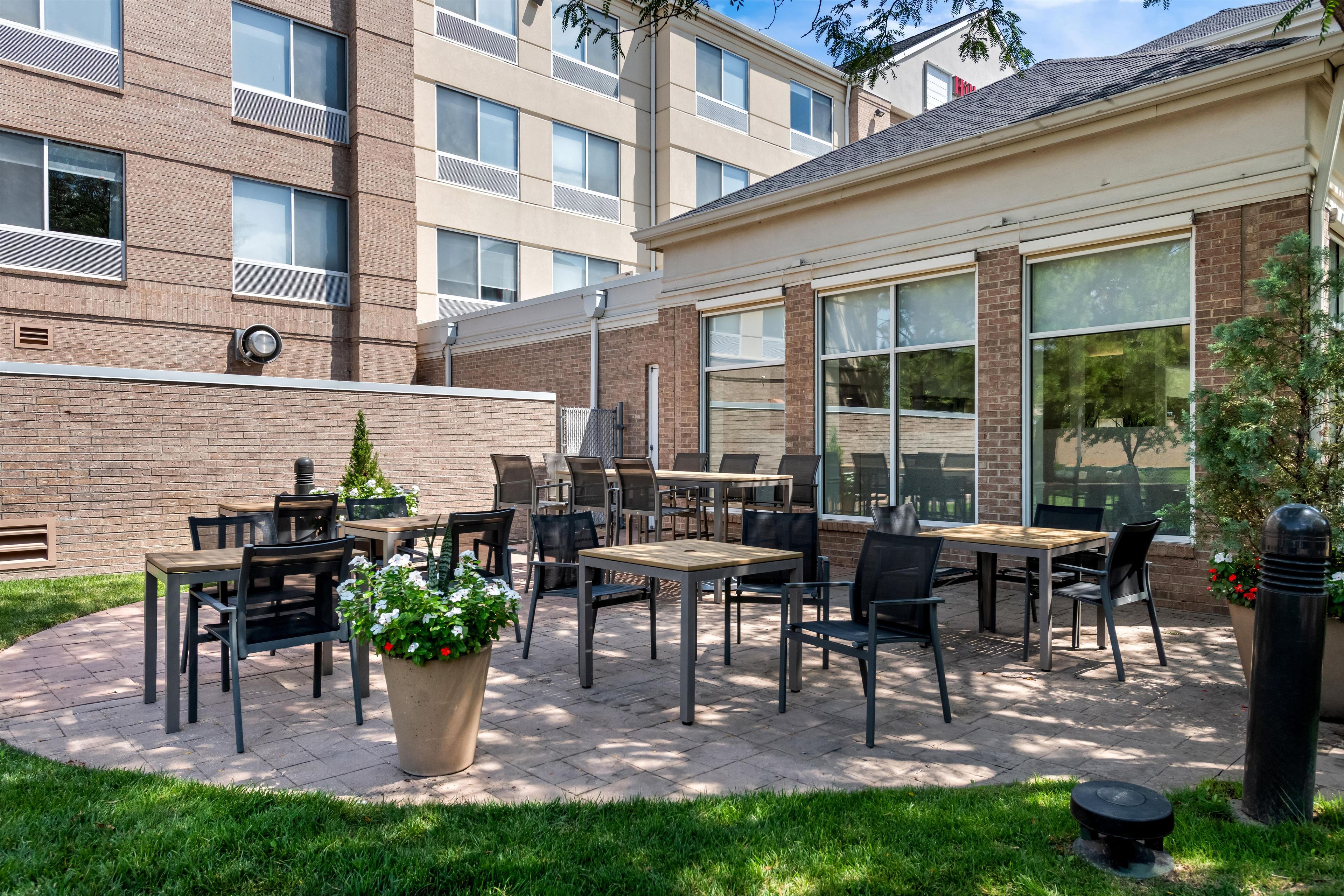 An outdoor patio area with tables and chairs, a potted plant, and a building with large windows in the background.