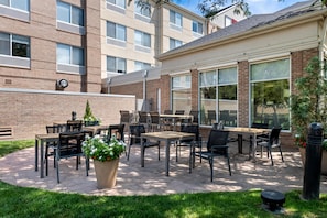Un espace extérieur aménagé en terrasse, avec des tables et des chaises, un pot de fleurs et, au fond, un bâtiment doté de grandes fenêtres.