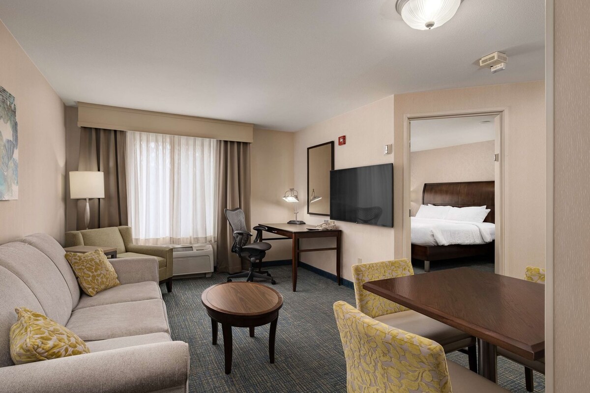 A hotel room with a sofa, a dining table, a desk with a chair, a bed, a TV, and a window with curtains.