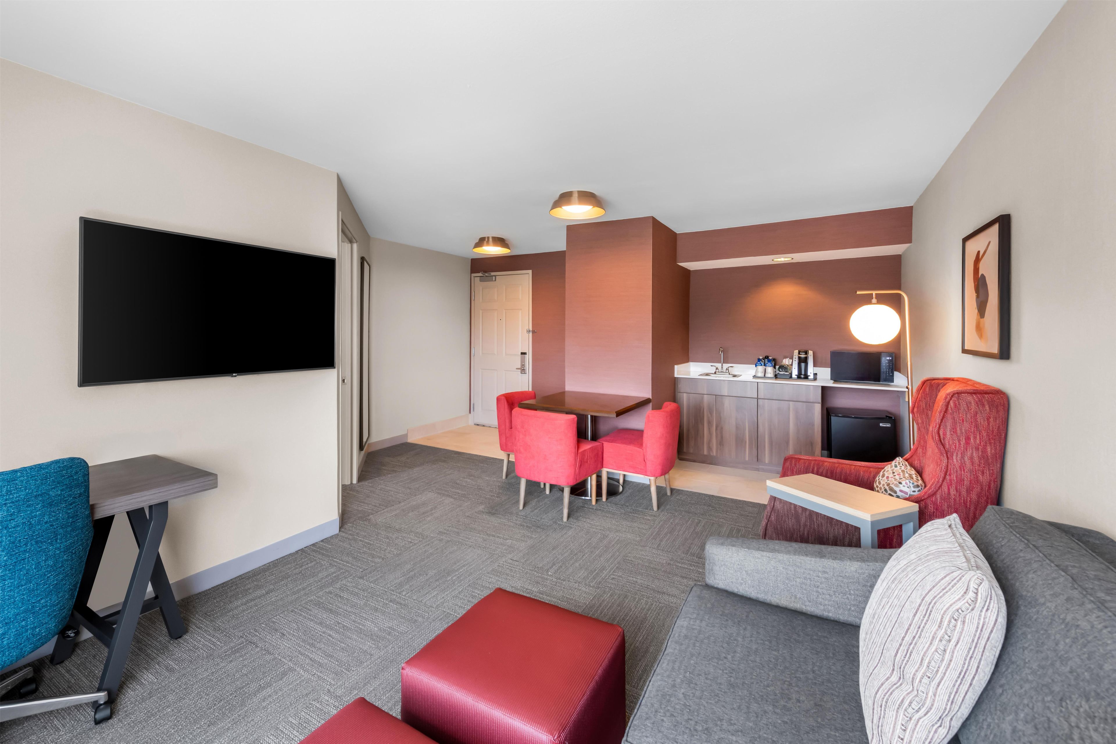 Ein Hotelzimmer mit Flachbildfernseher, einer Couch, einem roten Sessel, einem kleinen Tisch und einer Küchenzeile im Hintergrund.