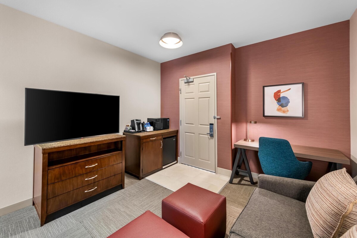 A modern hotel room with a flat-screen TV, wooden entertainment unit, kitchenette, seating area with a couch and armchair, and a door with a handle.