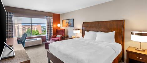 Habitación de hotel con una cama grande, un escritorio, una silla, una lámpara de escritorio, una mesita de noche, cortinas en la ventana, un equipo de aire acondicionado y un cuadro en la pared.