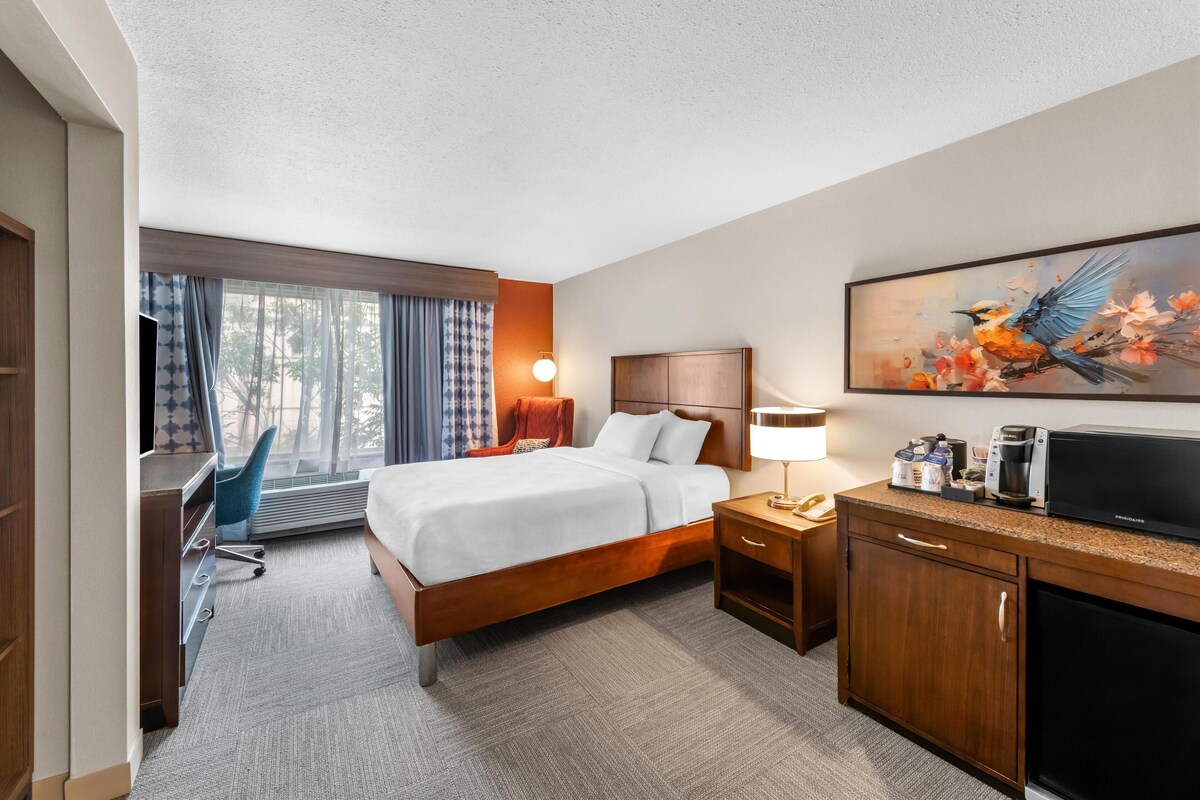 A hotel room with a bed, a desk, a chair, a TV, a coffee maker, and a painting on the wall.