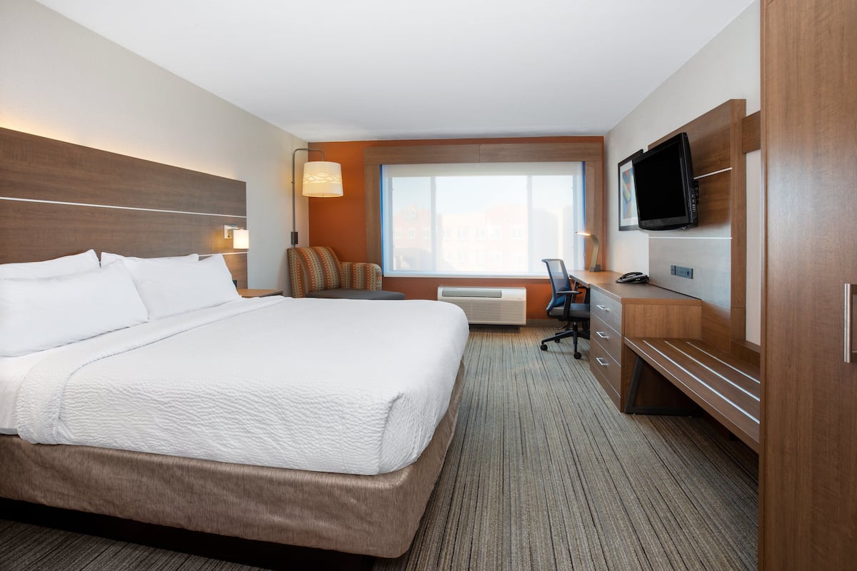 A hotel room with a large bed, a desk, a chair, a television, and a window with a view of buildings.