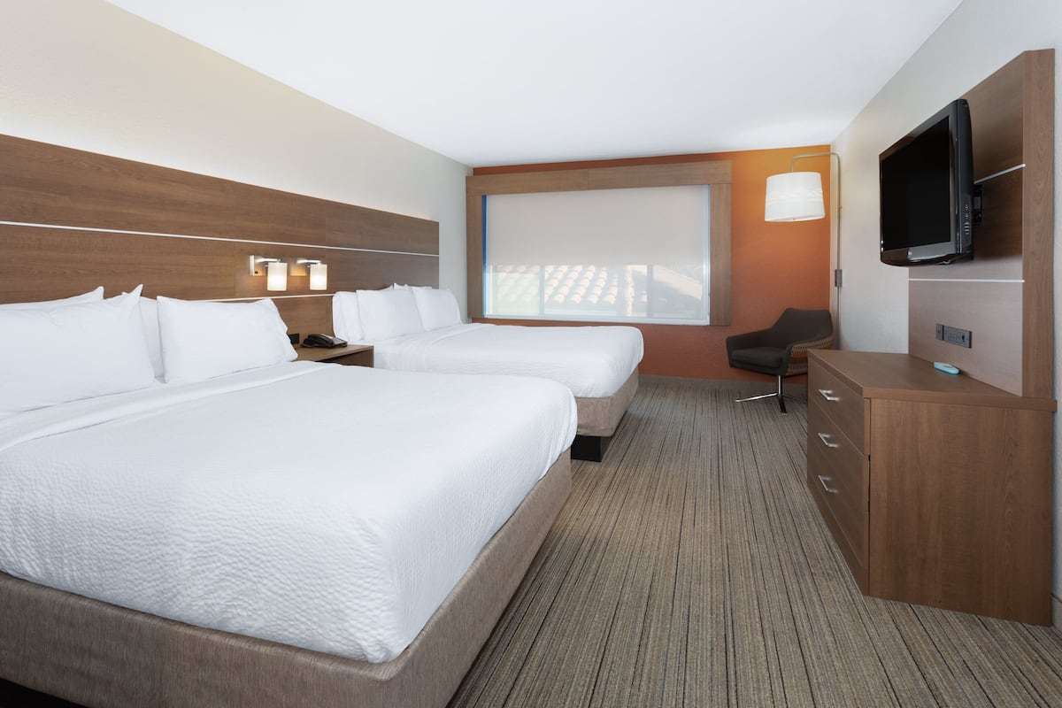 A hotel room with two beds, a desk, a chair, a TV, and a large window with blinds.