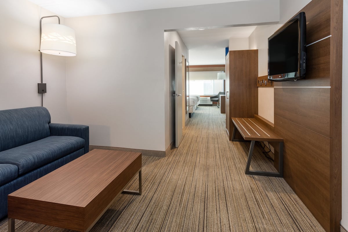 A hotel room with a blue sofa, a wooden coffee table, a TV mounted on a wooden wall, and a carpeted floor.