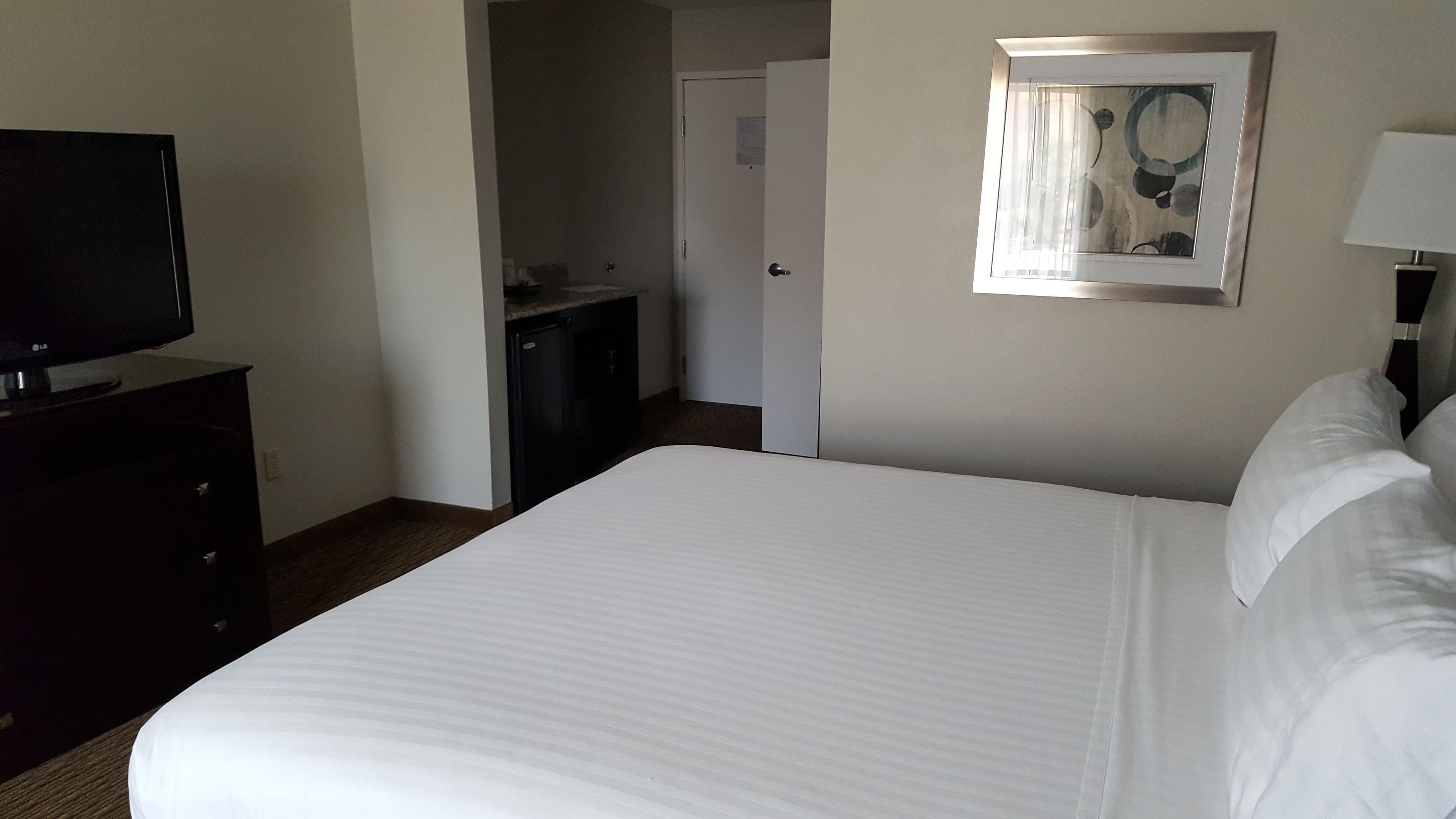 A hotel room with a large bed, a television on a dresser, a door leading to a bathroom, and a framed picture on the wall.