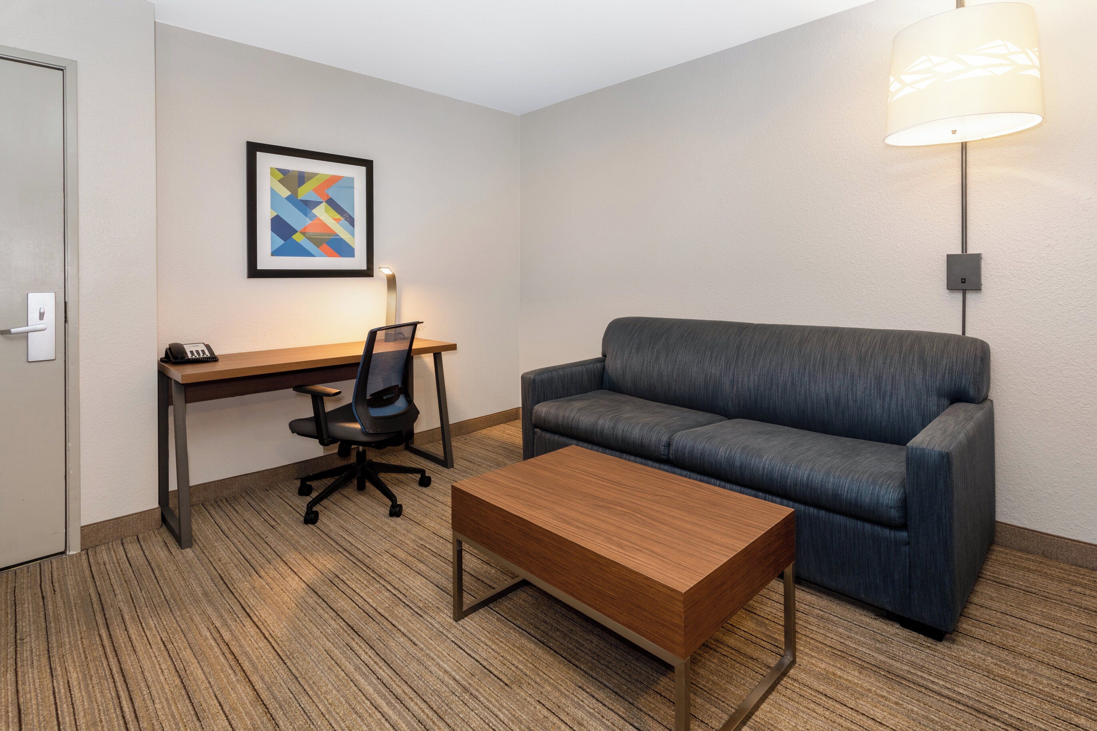 Une chambre d’hôtel avec un bureau en bois, un canapé bleu, une table basse en bois, un fauteuil de bureau noir et une œuvre d’art abstraite encadrée accrochée au mur.