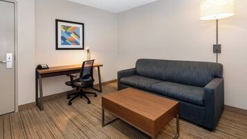 Une chambre d’hôtel avec un bureau en bois, un canapé bleu, une table basse en bois, un fauteuil de bureau noir et une œuvre d’art abstraite encadrée accrochée au mur.