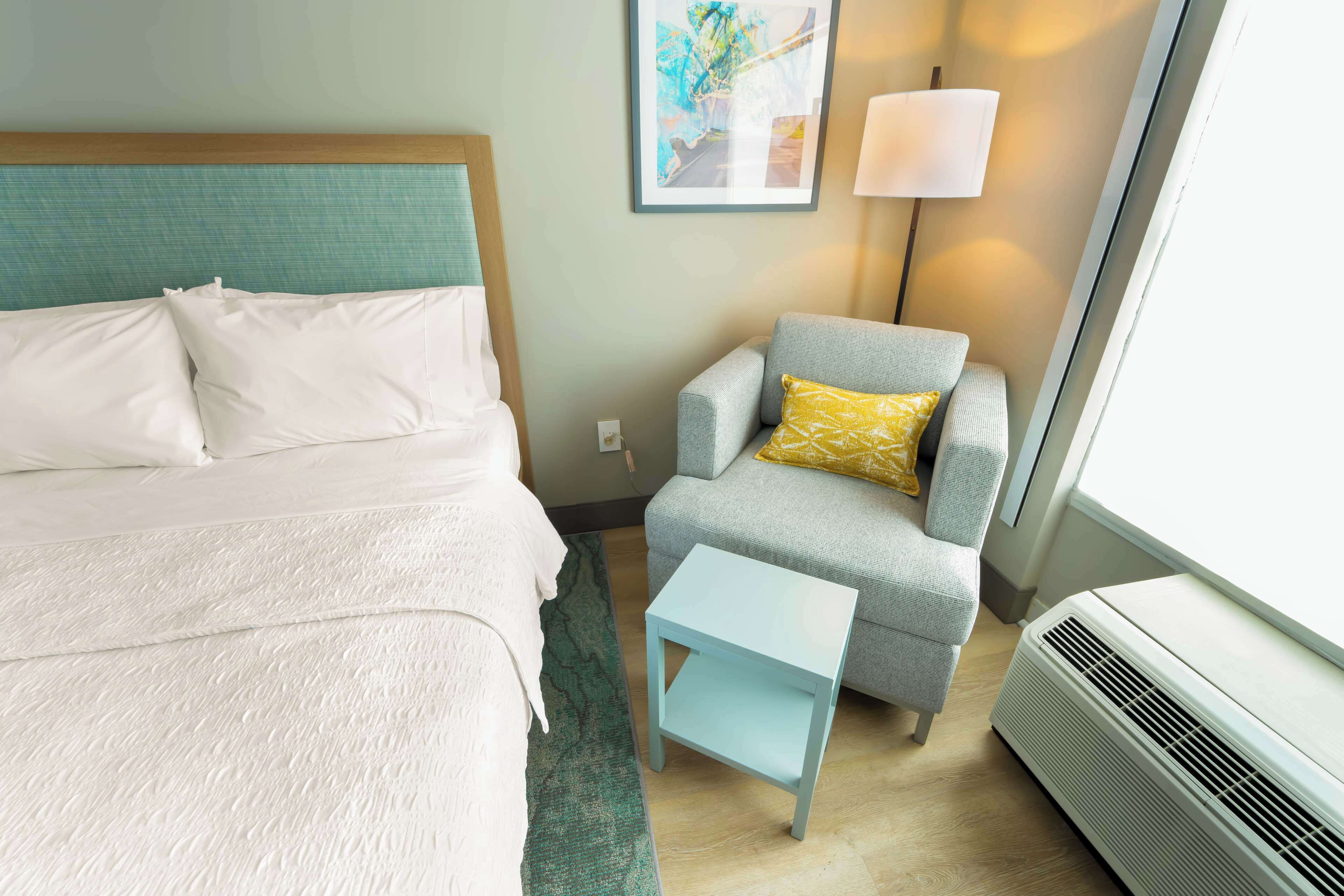 A hotel room with a bed, a chair, a small table, and an air conditioner.